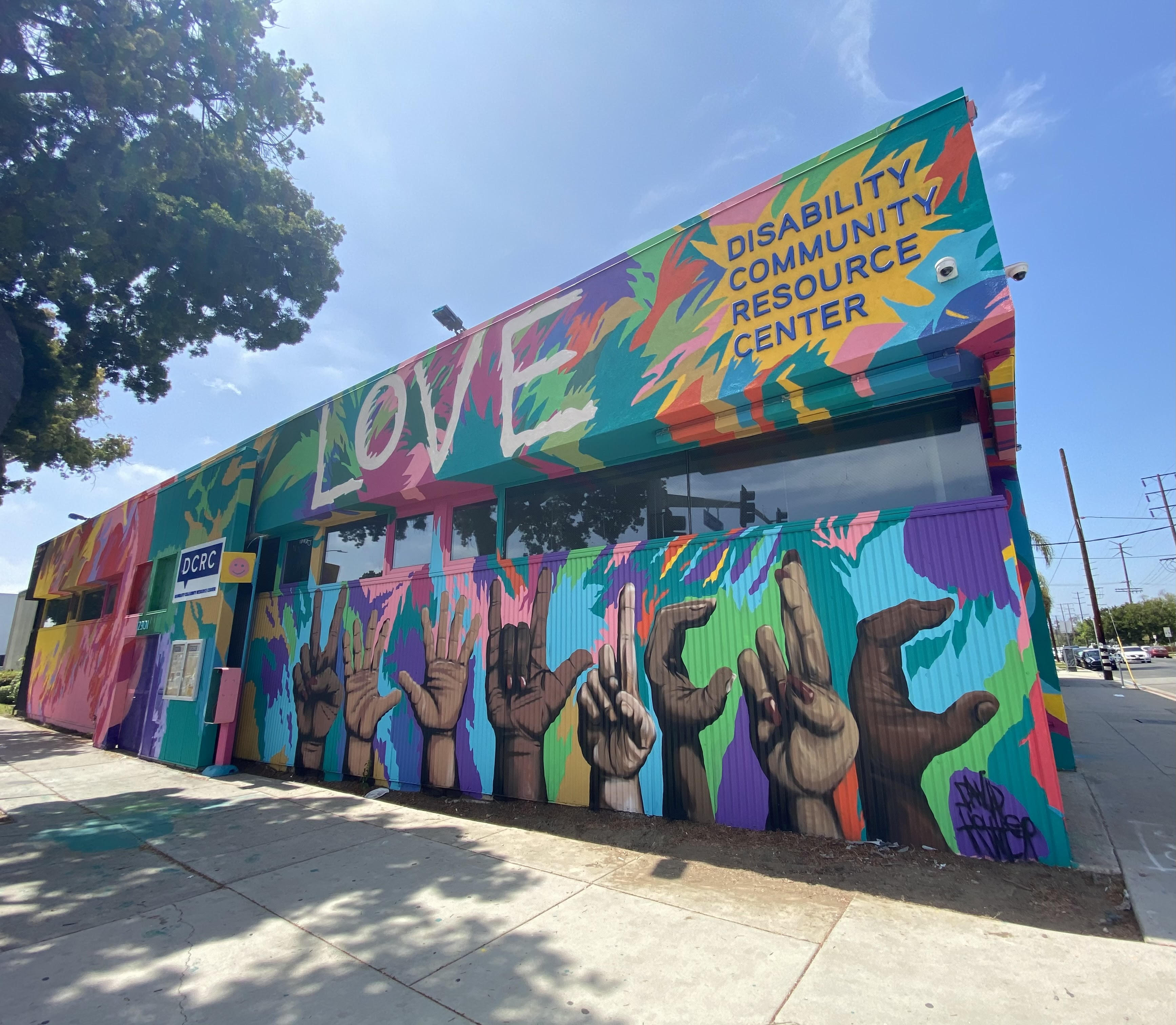 Image: Front of DCRC Building, with a colorful mural displaying the word Love on the top, the text of "Disability Community Resource Center", and Victory, Love, and the letters DCRC in American Sign Language